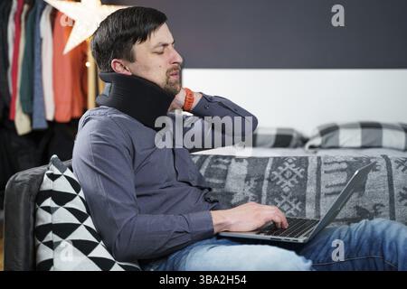 Tired strained man suffering from neck pain while working at computer. Male touchingly massages neck, puts on neck collar, sitting on couch, suffering Stock Photo