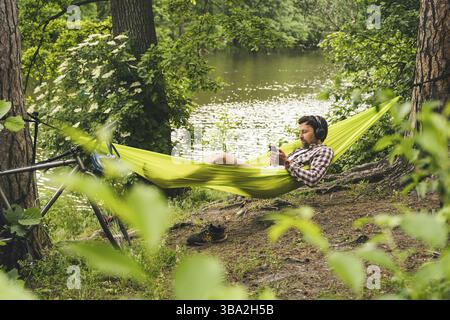 Young caucasian man resting in hammock, listening to music on headphones and using smartphone afterwards on bicycle in forest near the lake. Audio hea Stock Photo