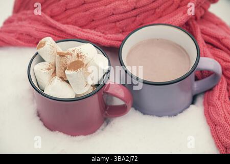 Hot chocolate with marshmallow in pink and violet two cups wrapped in a cozy winter pink scarf on the snow-covered table in the garden. Coloring and p Stock Photo