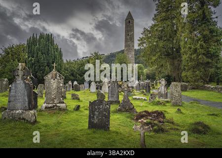 Wicklow, Ireland, August 2019 Medieval tower and ancient graves with Celtic crosses in Glendalough Cemetery. Moody storm sky in background, Europe Stock Photo