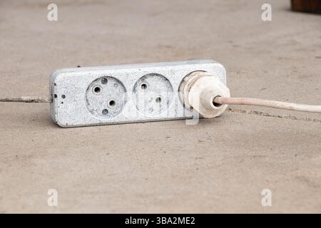 An outdoor extension cord with three sockets, white with black paint spots, lies on the concrete floor outside Stock Photo