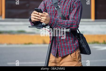 Young man wearing shirt leaning on electric scooter handlebar, holding mobile smartphone in his hands, closeup detail Stock Photo