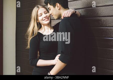 Portrait of attractive Mixed-race couple standing together against the background of a wooden wall. Dating, multiethnic wearing black clothes. Stylish Stock Photo