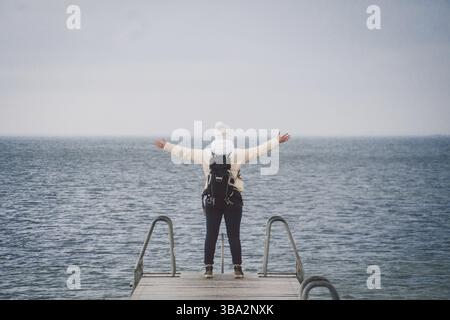 Concept of freedom relaxation. woman relaxing on pier. Backpacker hiker woman with backpack on sea wooden pier stands arms sideways looking to horizon Stock Photo
