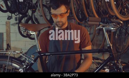 Theme small business bike repair. young Caucasian brunette man wearing safety goggles, gloves and apron uses hand tool repairing and adjusting crank a Stock Photo