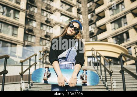 Stylishly dressed woman in blue denim jumpsuit posing with skateboard. Street photo. Portrait of girl holding skateboard. Lifestyle, youth concept. Le Stock Photo