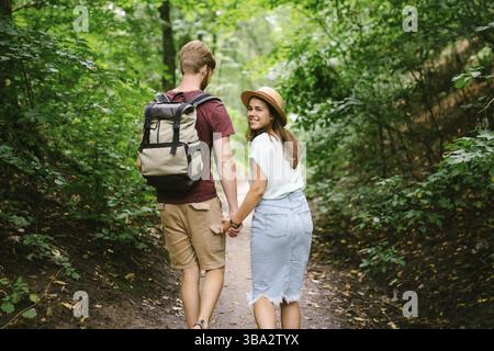 Happy caucasian couple are walking in a dense forest along the path holding hands, rear view. Hikers with backpack looking for place for picnic in woo Stock Photo