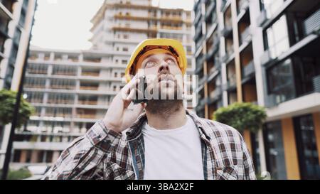 Portrait of young handyman making call while standing at construction area. Engineer talking on the phone on a construction site. Builder in helmet co Stock Photo