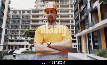 Young caucasian constructor with arms crossed in safety helmet and orange reflective uniform posing with proud expression at construction site. Proud Stock Photo