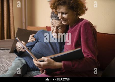 Senior woman and her adult daughter looking at photo album together on couch in living room, talking joyful discussing memories. Weekend with parents Stock Photo