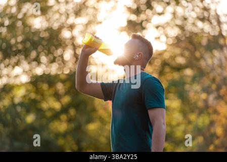 A sporty young man drinking water from a bottle after a run or workout in a summer park, active healthy lifestyle. Stock Photo