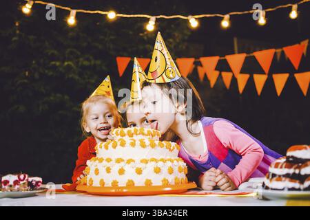 Children's birthday party. Three cheerful children girls eating cake with their hands and smearing their face. Fun and festive mood in the decorated c Stock Photo