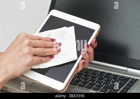 Woman cleaning tablet device with cotton paper tissue, wiping black screen, closeup detail pink color fingernails. Blurred laptop keyboard background Stock Photo
