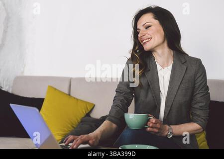 Middle age woman sitting in cafeteria holding coffee mug and working on laptop. Businesswoman checking email on laptop. Beautiful middle aged woman an Stock Photo