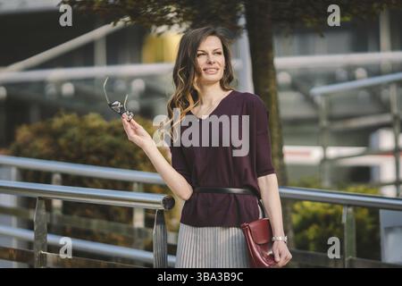 Caucasian adult lady woman in glasses posing near office building outside. Business lady in skirt holds hand glasses for vision bag in hand. Professio Stock Photo