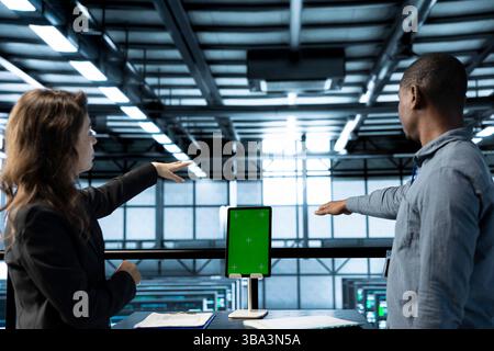 Computer scientists in data center use mockup tablet, inspecting server rigs, updating network topologies. Teamworking IT colleagues brainstorming, using chroma key device, ensuring stable operations Stock Photo