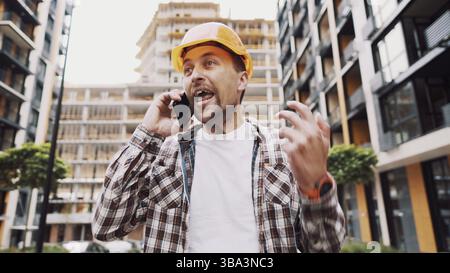 Angry builder, dissatisfied with deadline of work, swears at stress while talking to foreman on phone background of construction site. Wrathful archit Stock Photo