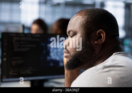 Tired man falling asleep while using artificial intelligence technology to optimize data center. Exhausted server hub expert taking break from using AI tech to streamline operations, slacking off Stock Photo