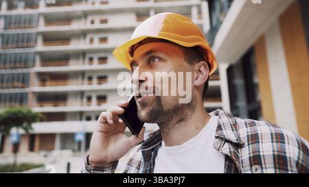 Portrait of young handyman making call while standing at construction area. Engineer talking on the phone on a construction site. Builder in helmet co Stock Photo