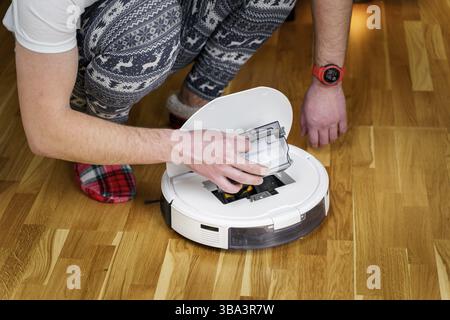 Caucasian young man in christmas pajamas doing vacuum cleaner robot maintenance. Male in winter patterned sleepwear and red socks slippers starts auto Stock Photo