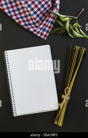 Blank cookbook with conventional and black uncooked Italian pasta Linguine, checkered napkin and basil on black stone background Stock Photo