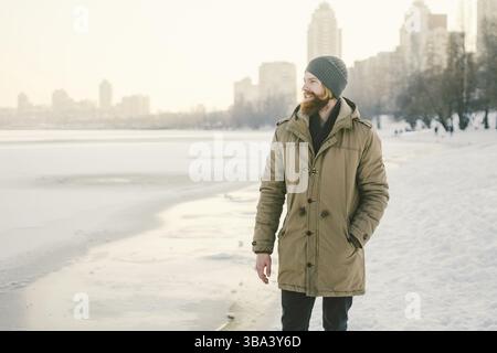 Close-up Caucasian young male red hair and beard in a hat and a park coat posing winter model against a background of a lake to freeze snow Stock Photo