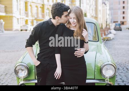 Young multiracial couple, male and female lovers heterosexual people students. Beautiful models posing standing near a retro car in the city. Dressed Stock Photo