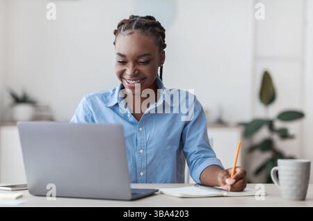 Online Education. Young Black Female Student Using Laptop At Home, Taking Notes Stock Photo