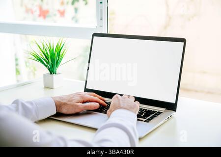Over shoulder view of a young business man using computer laptop in front of an blank white computer screen in his home living room. Photo ready for m Stock Photo
