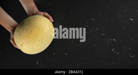 Top down view, woman hands holding cantaloupe melon over black marble like board. Wide banner with space for text on right side, Liptovsky Hradok, Slo Stock Photo