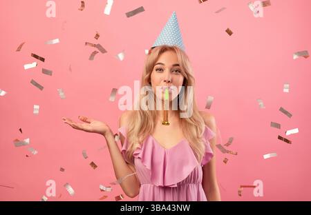 Gorgeous young woman in birthday cap blowing party horn on pink studio background with falling confetti Stock Photo