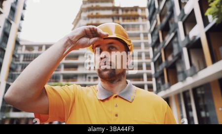 Serious Caucasian builder stands at construction site puts on orange hard hat. Safety first of all, construction profession. Low angle shot. Foreman i Stock Photo