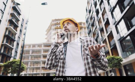 Angry male architect in orange hard hat talks swears on phone at construction site. Builder in plaid shirt emotionally displeased negotiates by cell p Stock Photo