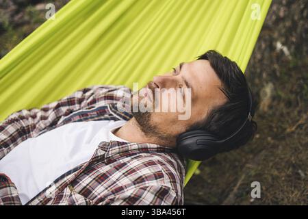 Young caucasian man resting in hammock, listening to music on headphones and using smartphone afterwards on bicycle in forest near the lake. Audio hea Stock Photo