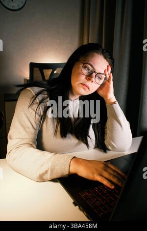 Woman working on computer late evening at home office. Concentrated female freelancer trying to solve business problems. Working and suffering from we Stock Photo