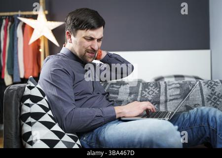 Man employee after accident working at home behind laptop and feels pain in neck, uses black neck collar support. Male with neck injury freelance prog Stock Photo