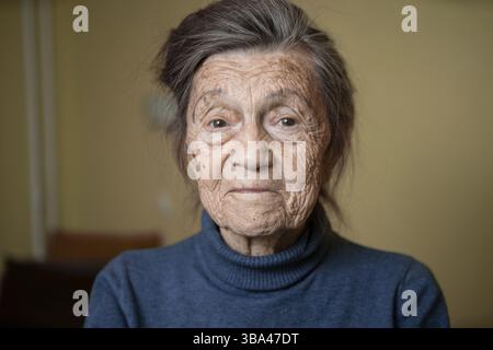 90 year old cute elderly woman with gray hair and wrinkles face, wearing sweater, portrait large, smiling and looking joyfully, background of room. Th Stock Photo