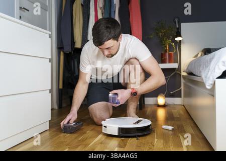 Caucasian young man doing white robot vacuum cleaner maintenance. Autonomous wireless modern vacuum cleaner. Male repairing robot cleaner home on floo Stock Photo