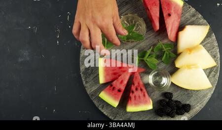 MELON WATERMELON BLACKBERRY AND MINT SHOT OF THE HAND ON A DARK BACKGROUND. RIPE CHUNKS OF WATERMELON JUICY MELON BLACKBERRY MINT WITH GLASS AND HAND Stock Photo