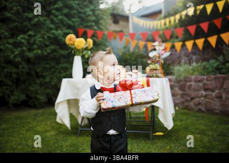 Surprise for the birthday. the boy is holding a box with a gift in the yard on the background of a festive table with a cake. dressed in a black suit Stock Photo