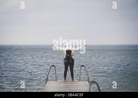 Concept of freedom relaxation. woman relaxing on pier. Backpacker hiker woman with backpack on sea wooden pier stands arms sideways looking to horizon Stock Photo