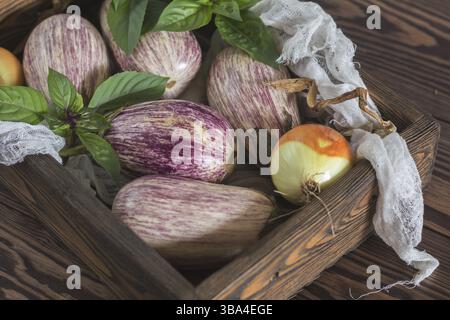 Purple graffiti eggplants, onion and green fresh basil in a wooden box in a vintage wooden background in rustic style, selective focus Stock Photo