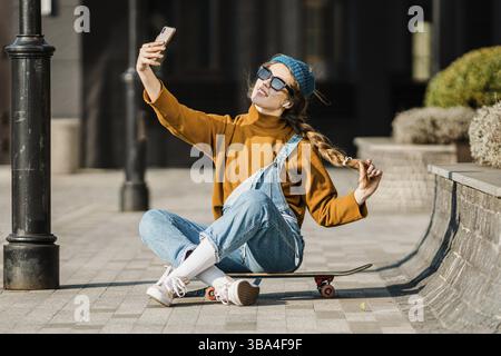Young woman sitting on skateboard, using phone. skateboarding woman in city. female skate boarder with smartphone and skateboard. cute skater sitting Stock Photo