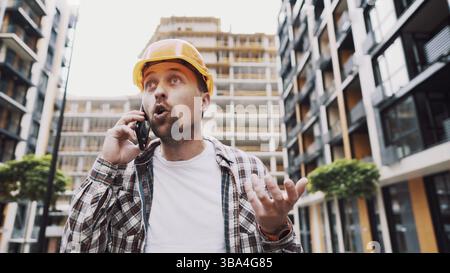Angry builder, dissatisfied with deadline of work, swears at stress while talking to foreman on phone background of construction site. Wrathful archit Stock Photo