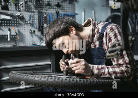 Male mechanic working in bicycle repair shop, mechanic repairing bike using special tool, wearing protective gloves. Young attractive serviceman fixin Stock Photo