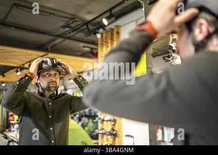 Young man came to the bicycle store. He is measuring the helmet. Male chooses helmet in sports equipment store. Purchase of new sports helmet. Custome Stock Photo
