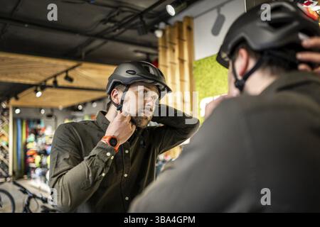 Caucasian man trying on black bicycle helmet near mirror in sporting goods store. Male buyer chooses safety helmet for cycling. Shopping in bicycle st Stock Photo