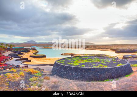 Lago de la Santa in Lanzarote features volcanic rock formations and calm saltwater pools under a cloudy sky. Circular stone structures with green moss Stock Photo