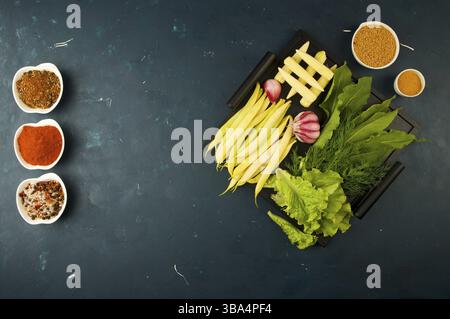 THE VEGETABLES IN THE BOX ON THE STONE A DARK BACKGROUND. YOUNG GREENS ONION GARLIC ZUCCHINI BRIGHT SPICES LAY IN A WOODEN TRAY WITH HANDLES ON A DARK Stock Photo