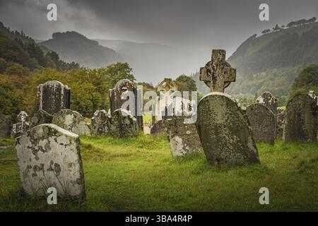 Ancient graves with Celtic crosses in Glendalough Cemetery. Autumn forest, mountains, rain and dramatic storm sky in background, Wicklow, Ireland, Eur Stock Photo